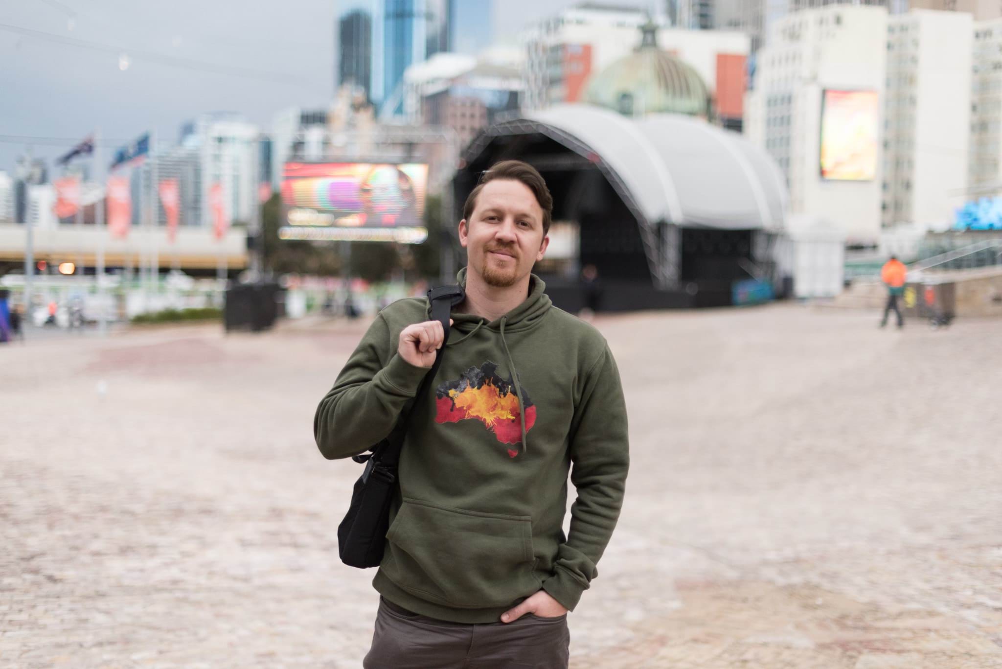 A First Nations man stands in the middle of Federation Square in Melbourne wearing a hoodie which shows the map of Australia decorated in the colours of the Aboriginal flag.